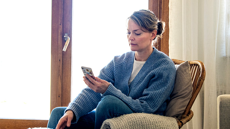 Medelålders kvinna sitter i fåtölj framför fönstret och chattar med 1177 direkt via sin mobiltelefon